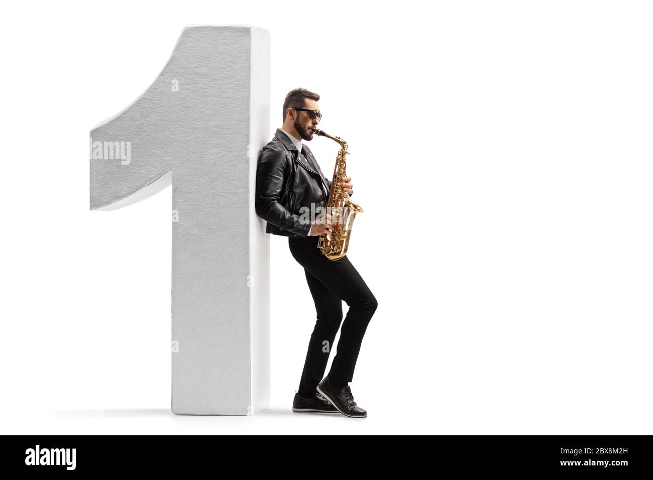 Full length profile shot of a young male musician in a leather jacket playing a saxophone and leaning on number one isolated on white background Stock Photo