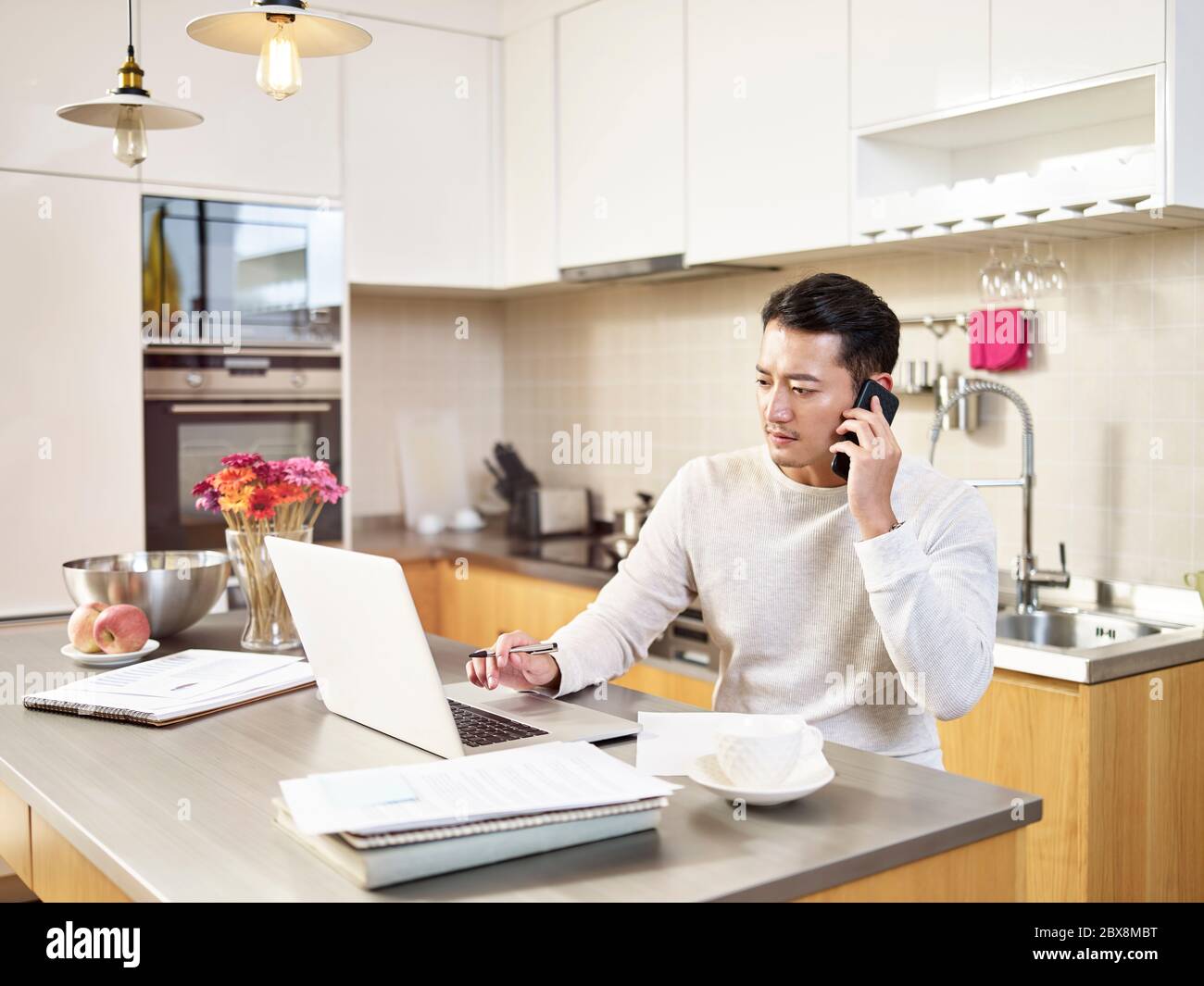 young asian business man sitting kitchen counter working at home looking at laptop computer talking on mobile phone Stock Photo