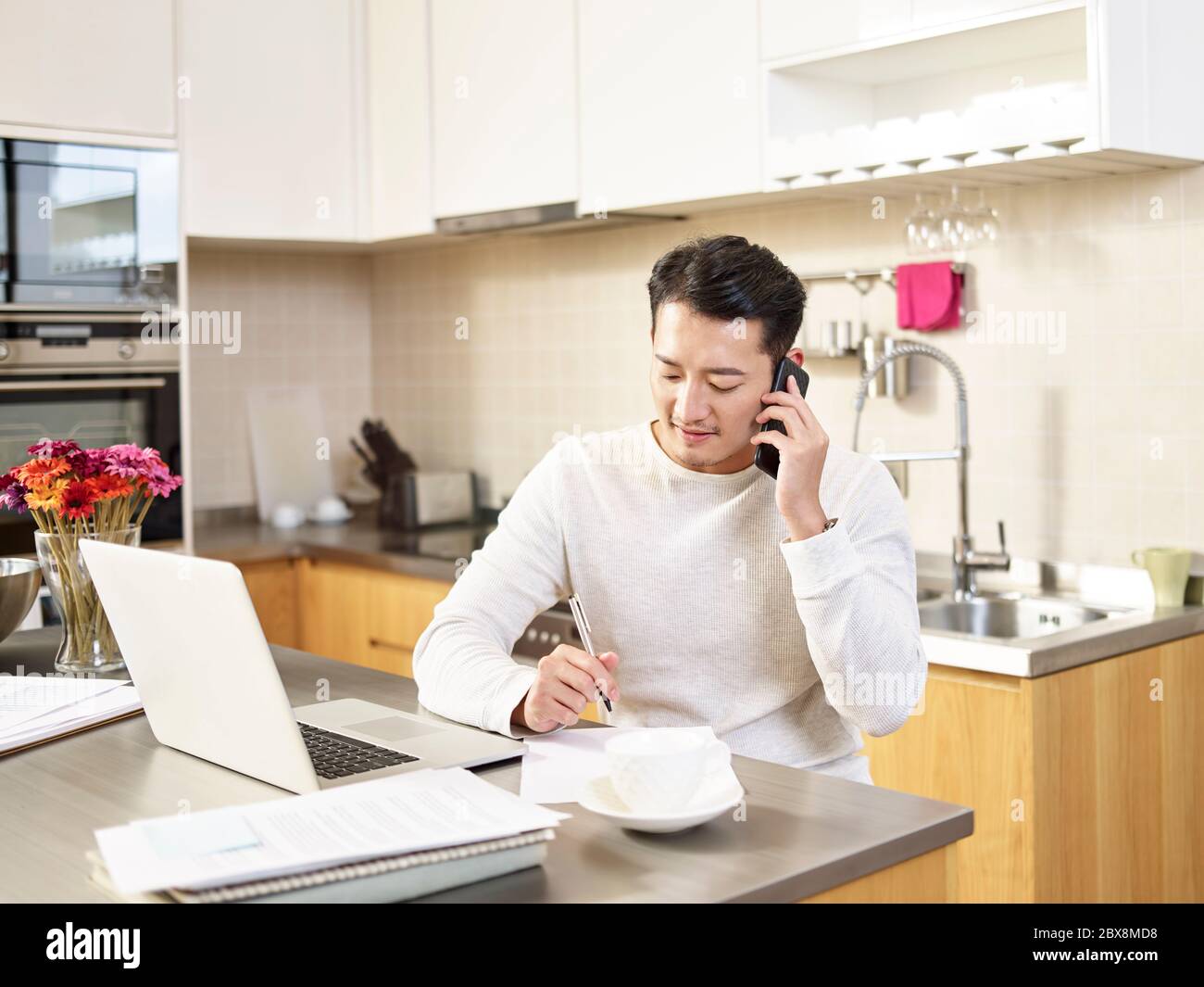 young asian business man sitting kitchen counter working at home talking on cellphone taking notes Stock Photo