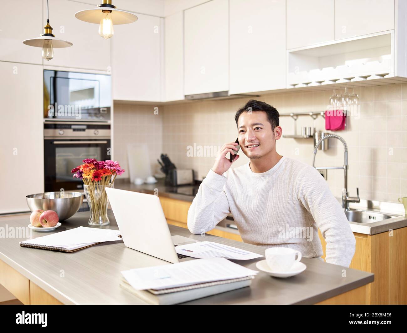 young asian business man sitting kitchen counter working at home talking on mobile phone Stock Photo