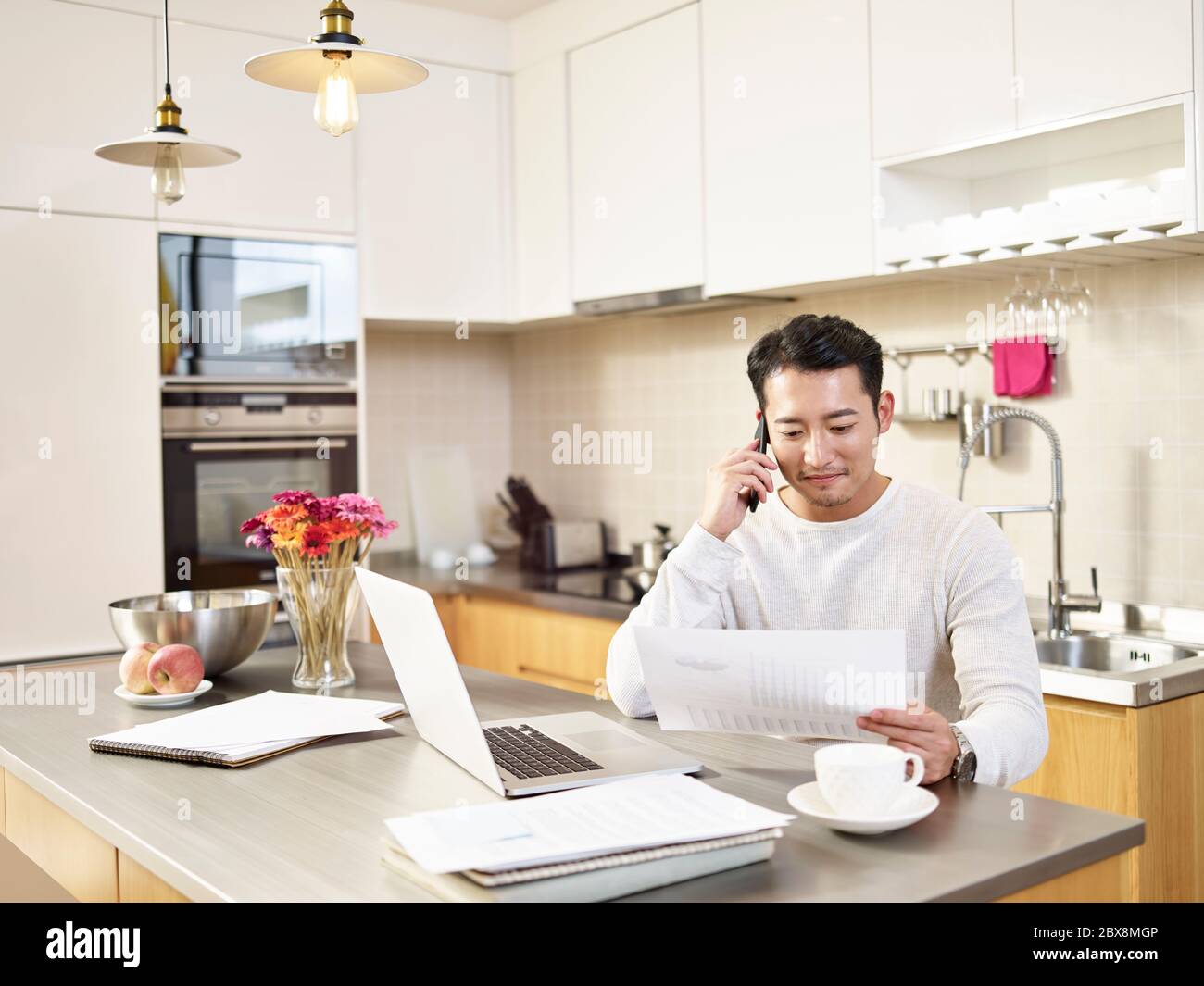 young asian business man sitting kitchen counter working at home looking at report talking on cellphone Stock Photo