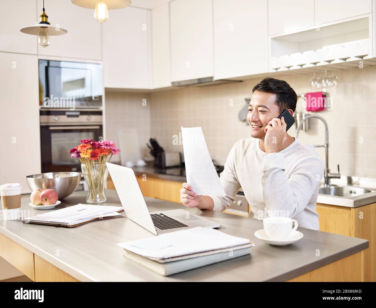 young asian business man sitting kitchen counter working at home looking at report talking on cellphone Stock Photo