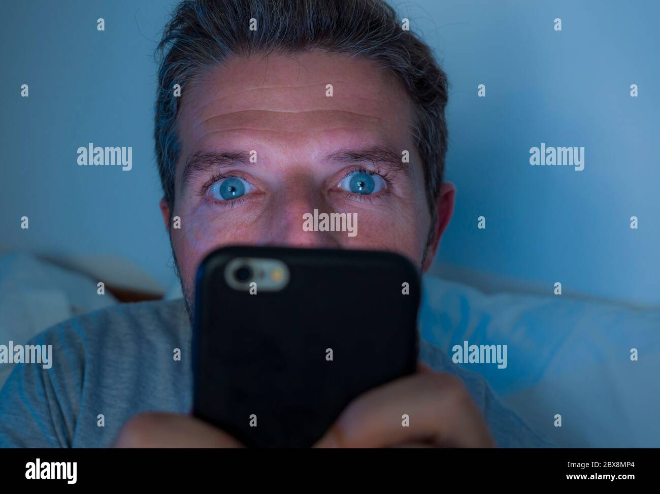 young attractive and crazy internet addict man with blue eyes lying on bed late at night in dark and dim light networking on mobile phone or online da Stock Photo