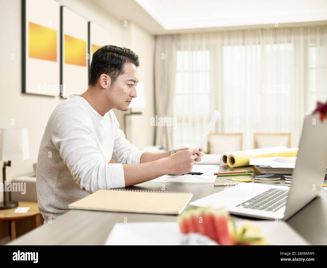 young asian man design professional working from home sitting at kitchen counter reading a document (artwork in background digitally altered) Stock Photo