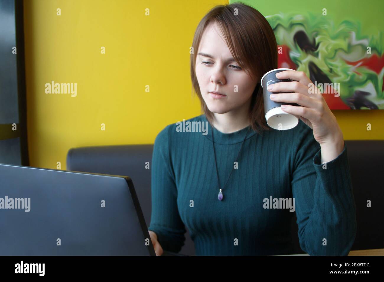 A girl with a pensive calm expression on her face sits at a laptop at a table in a cafe on a brown sofa against a yellow wall. He is looking at a computer monitor, holding a paper cup with coffee in his hand. Stock Photo
