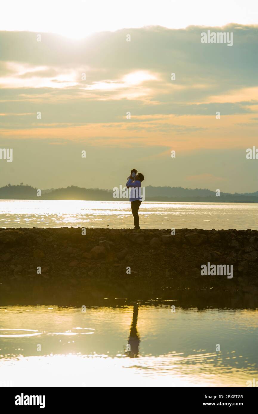 Silhouette of young happy man with his little son or daughter in his arms , the father raising up the baby in front of the sea at beach boardwalk enjo Stock Photo