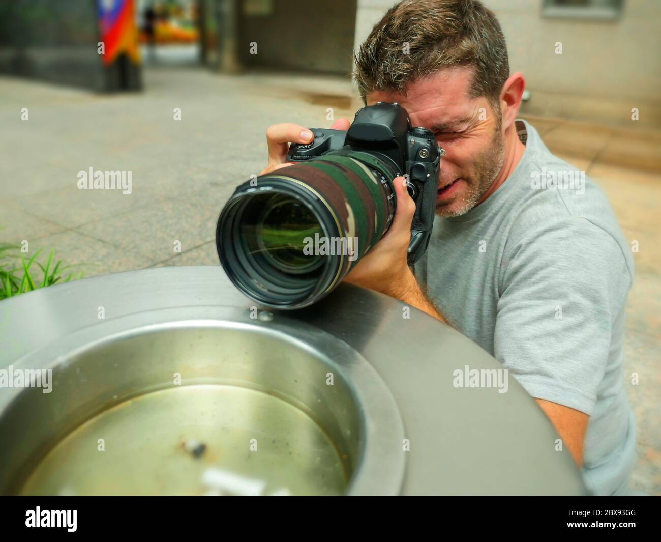 urban background lifestyle funny portrait of young paparazzi photographer man in action hidden behind city paper basket stalking for shooting exclusiv Stock Photo
