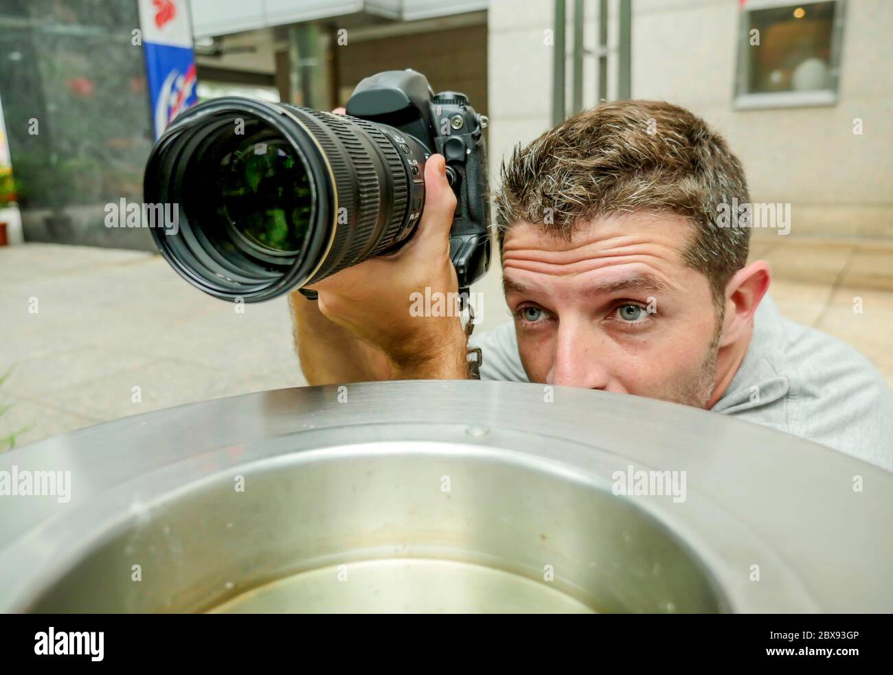 urban background lifestyle funny portrait of young paparazzi photographer man in action hidden behind city paper basket stalking for shooting exclusiv Stock Photo