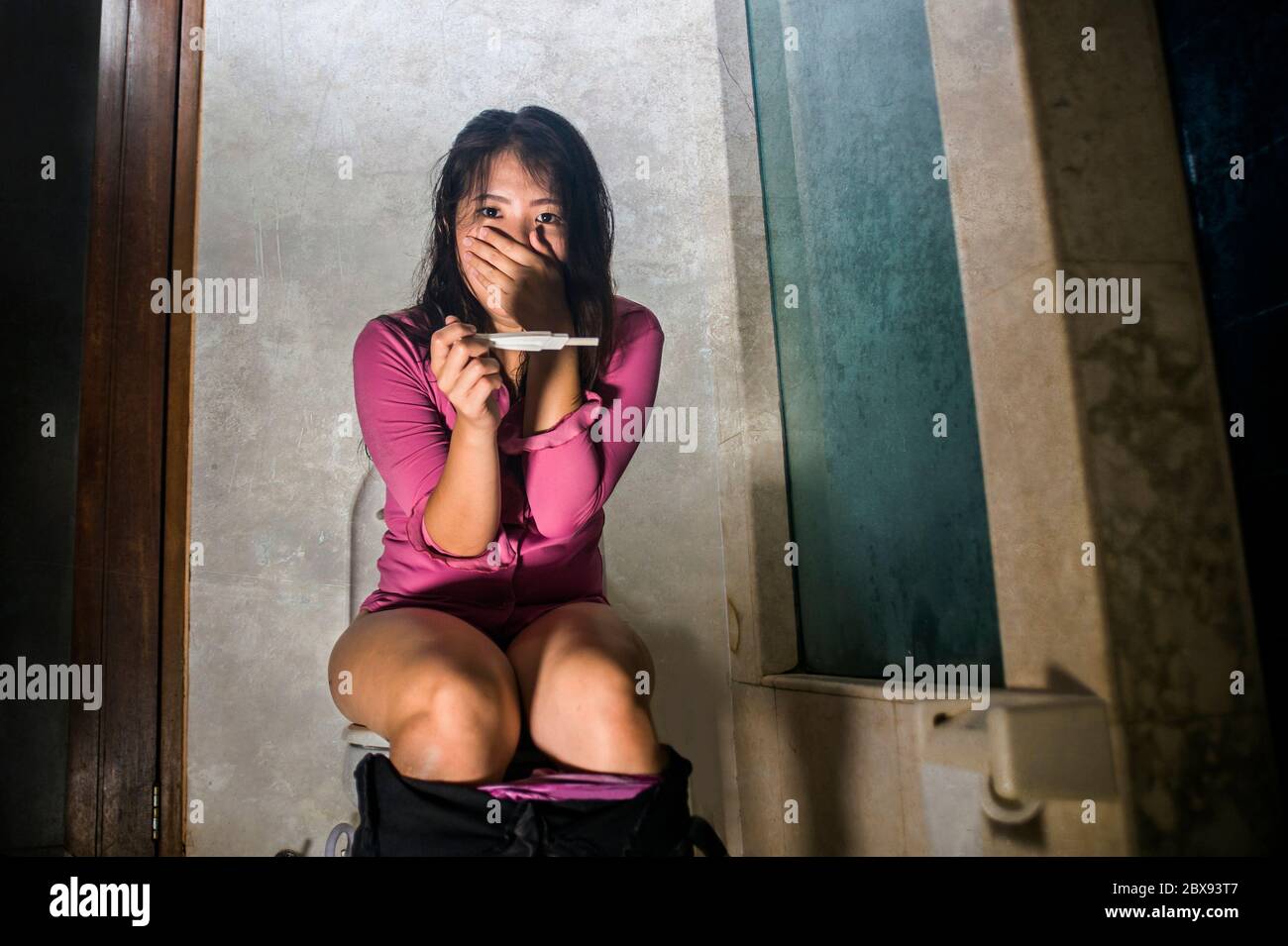 young scared and desperate pregnant Asian Japanese woman or teenager checking positive result on predictor test sitting on toilet WC crying overwhelme Stock Photo