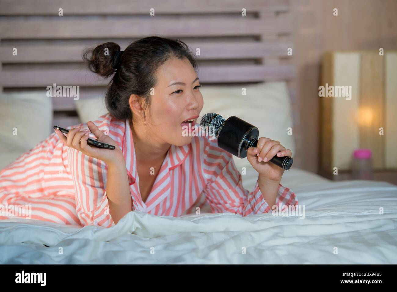 Young happy and beautiful Asian Korean teenager girl singing karaoke song excited at home bedroom holding mobile phone playing on bed excited and chee Stock Photo