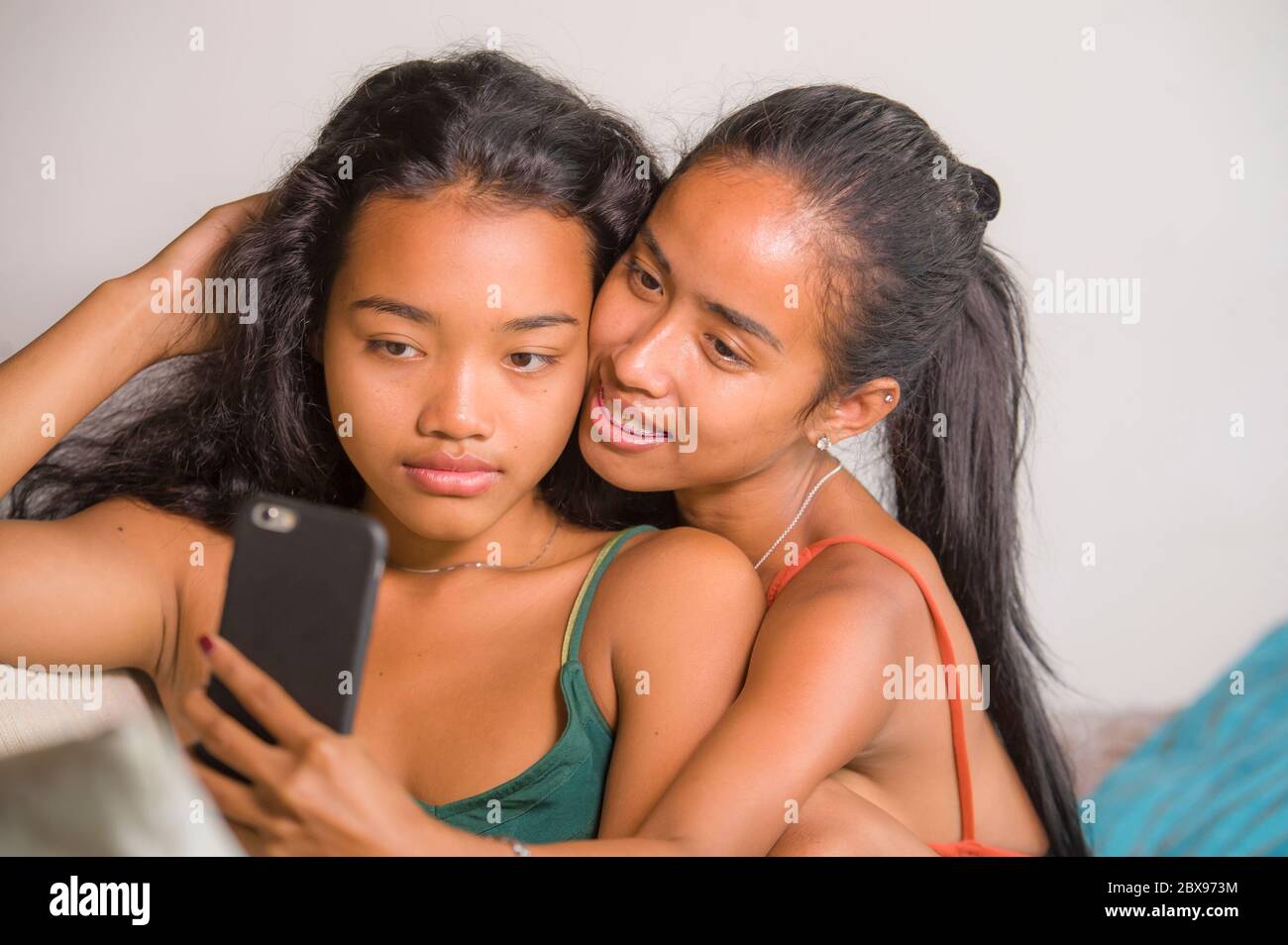 young happy and beautiful Asian sisters or girlfriends couple smiling cheerful taking selfie photo with mobile phone at home couch playful laughing to Stock Photo picture