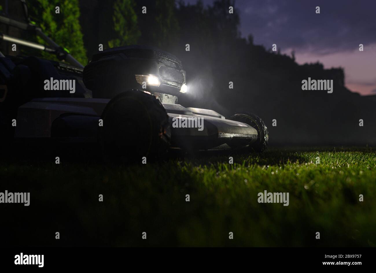 Backyard Grass Mowing During Late Summer Evening Hours. Modern Electric Grass Mower with LED Head Lights. Stock Photo
