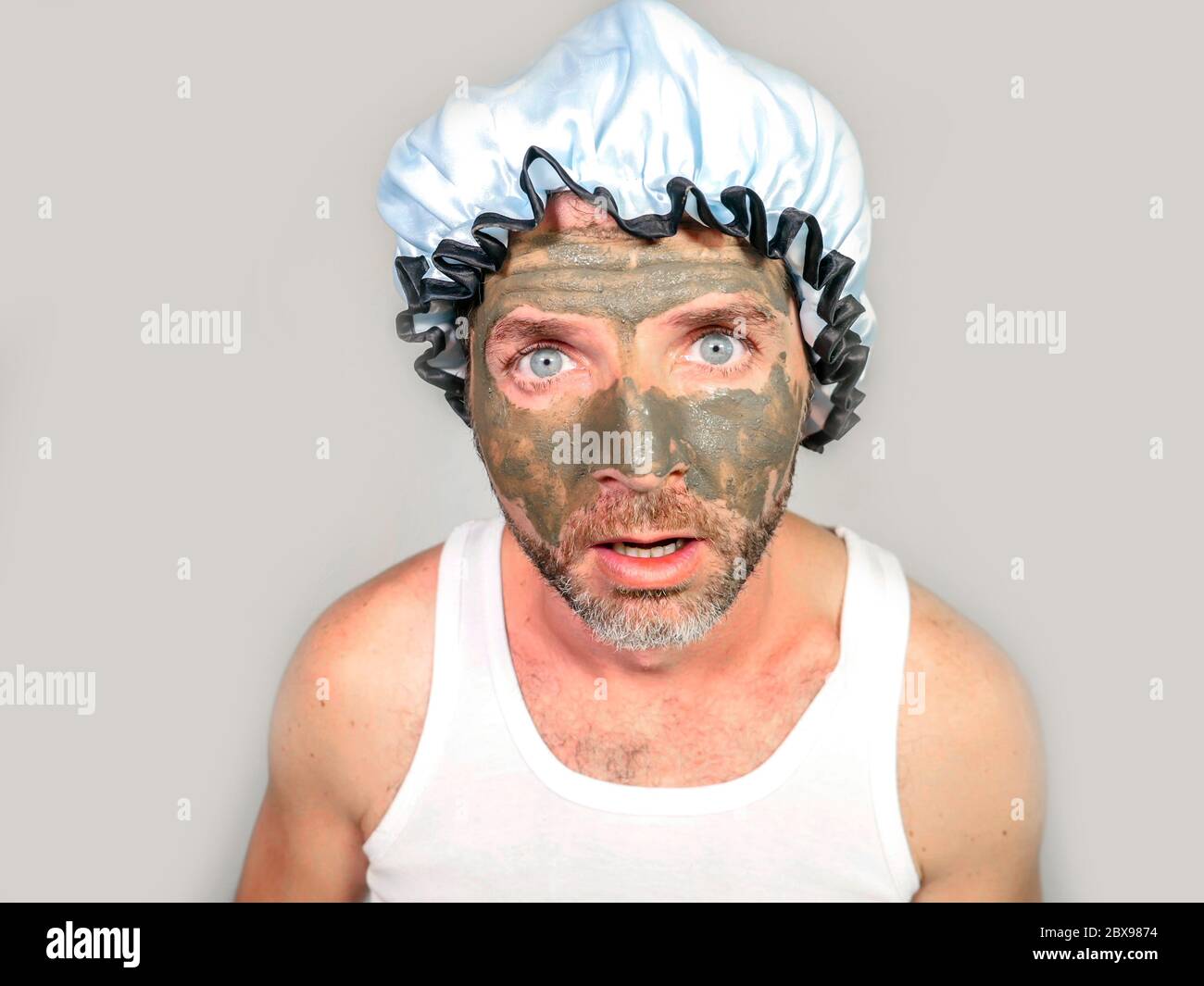 lifestyle funny portrait of young messy and weird looking man with shower cap and cream on his face horrified seeing himself ugly on bathroom mirror a Stock Photo