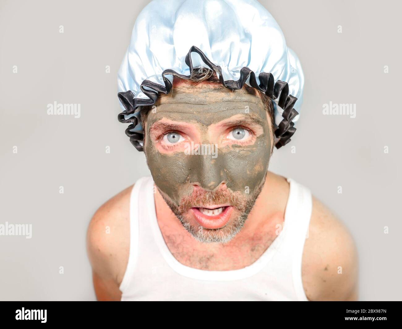 lifestyle funny portrait of young messy and weird looking man with shower cap and cream on his face horrified seeing himself ugly on bathroom mirror a Stock Photo