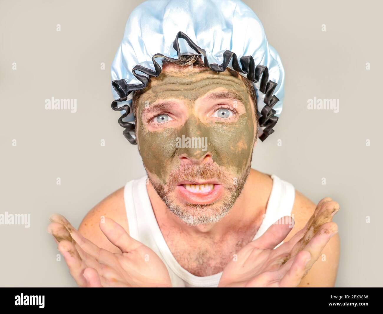 lifestyle funny portrait of young messy and weird looking man with shower cap and cream on his face horrified seeing himself ugly on bathroom mirror a Stock Photo