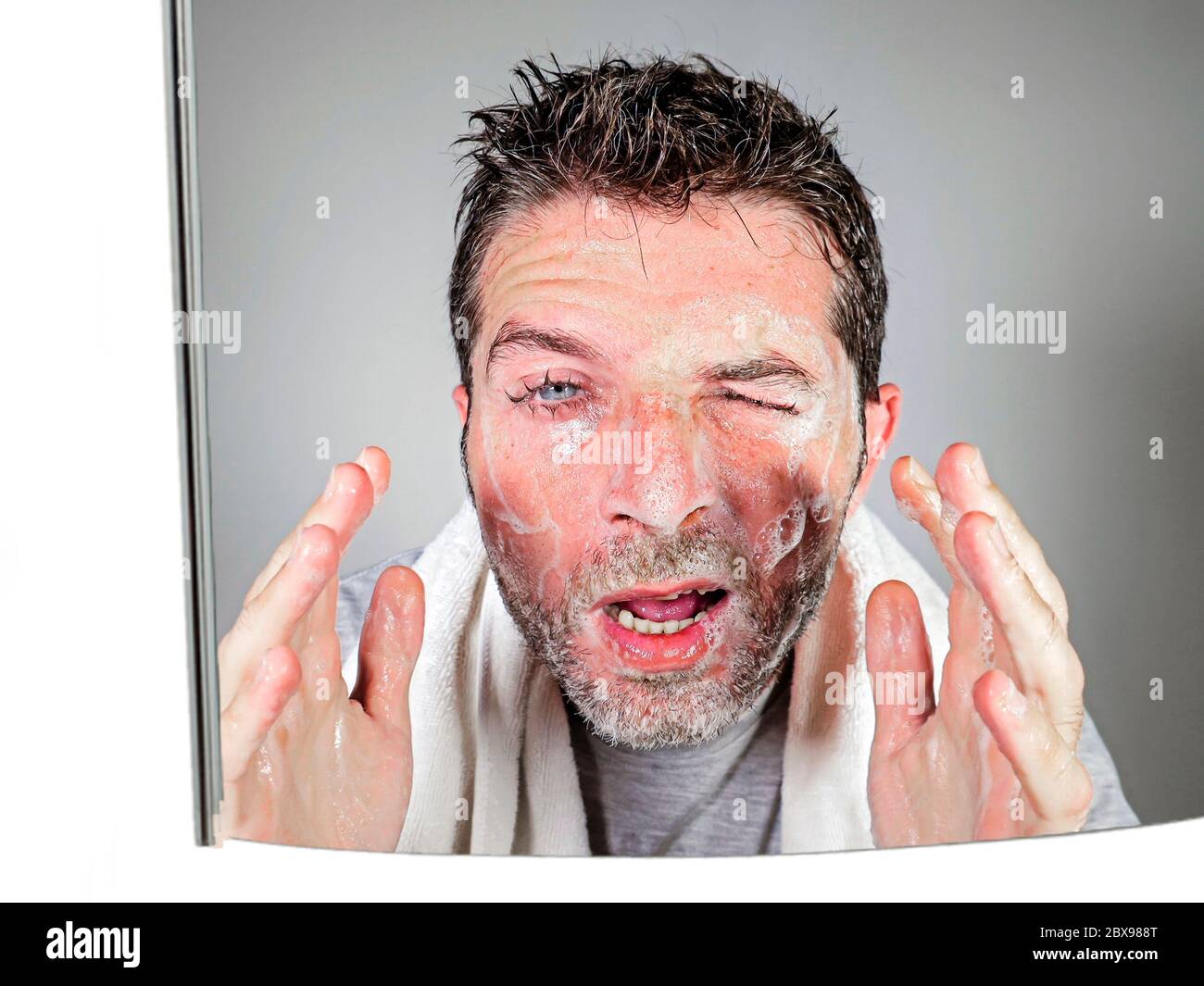mirror reflection portrait of young attractive and funny man washing face with soap with itchy