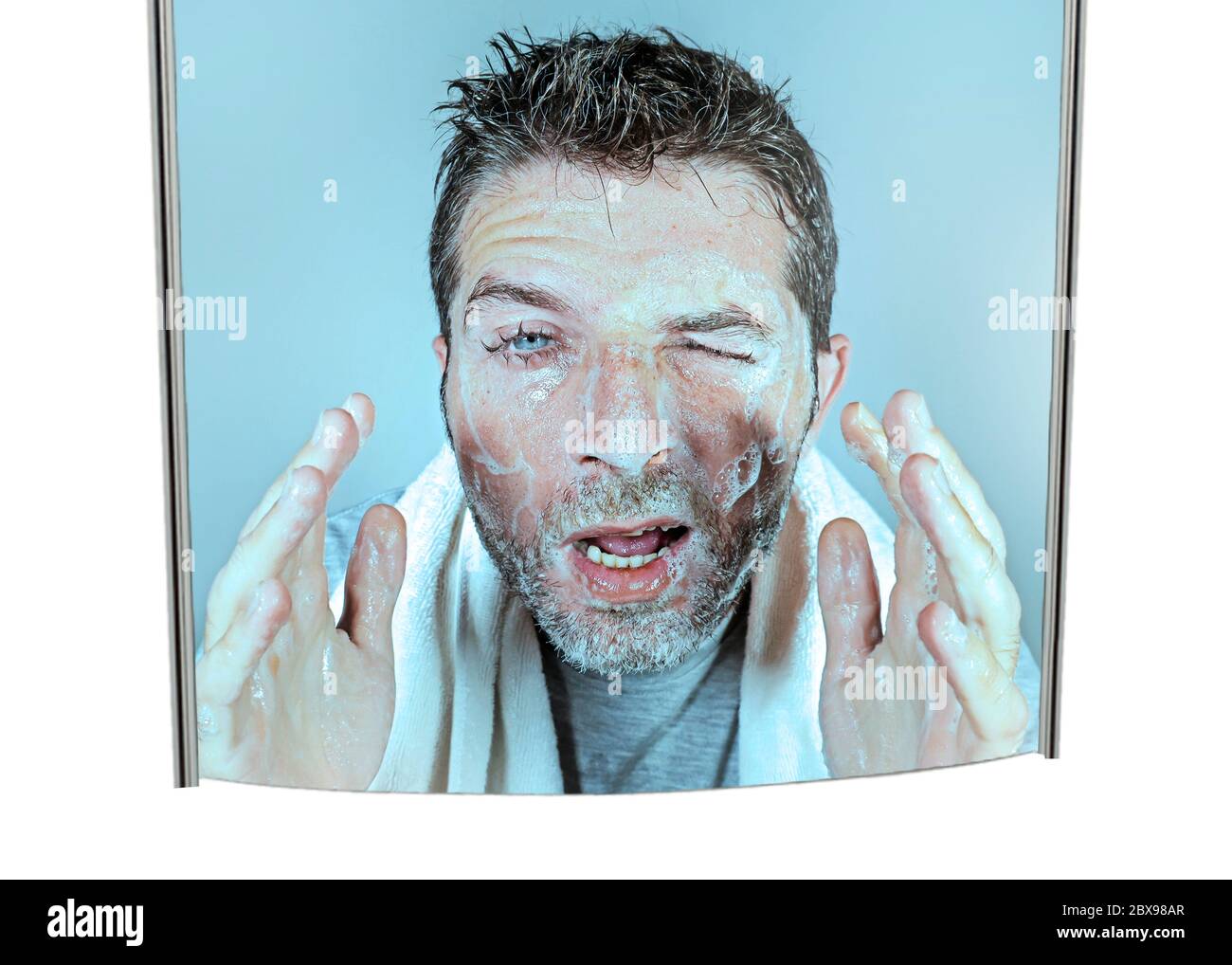 mirror reflection portrait of young attractive and funny man washing face with soap with itchy eyes and messy face expression applying beauty facial s Stock Photo