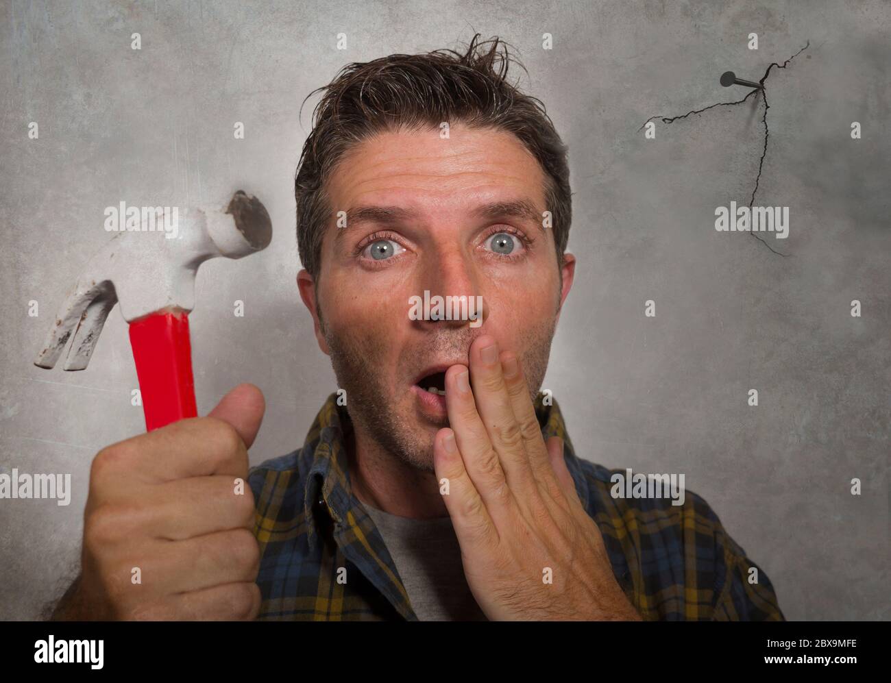 funny portrait of man holding hammer driving a nail for hanging a frame but making a mess cracking the wall as a disaster DIY guy and messy domestic r Stock Photo