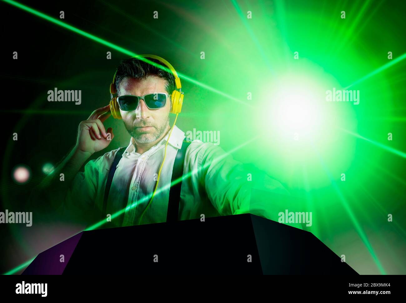young attractive and cool DJ in shirt and suspenders remixing music at night club using headphones in party strobo and laser lights background in club Stock Photo