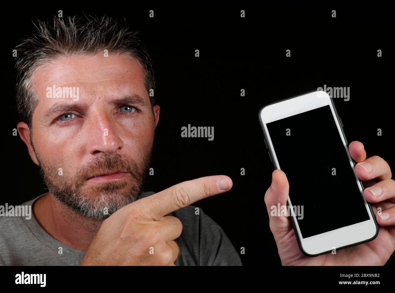 young handsome and attractive Caucasian man holding mobile phone in his hand pointing it with the finger isolated on black background in internet tech Stock Photo