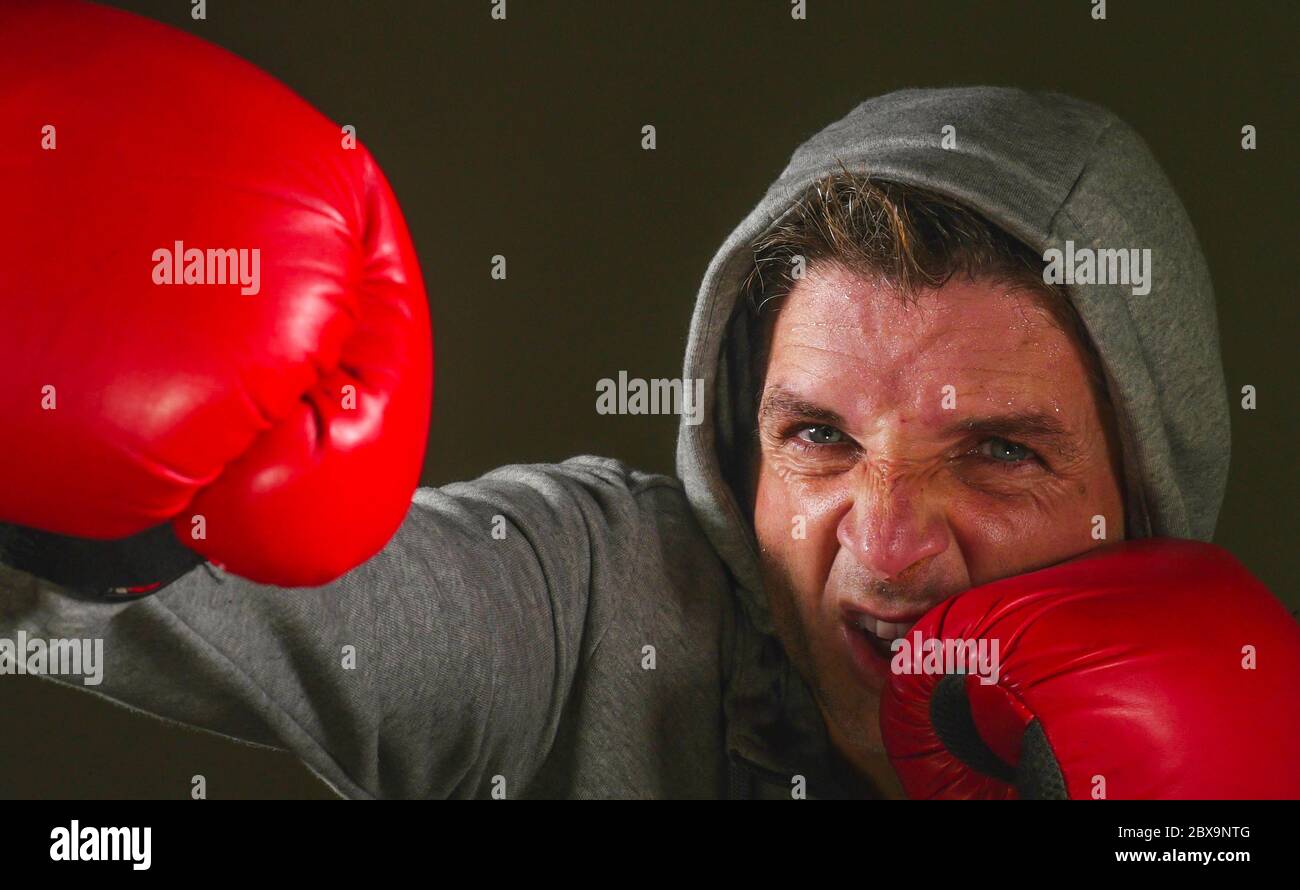 close up face portrait of young attractive and fierce looking man in boxing gloves throwing punch aggressive isolated on dark background in sport and Stock Photo