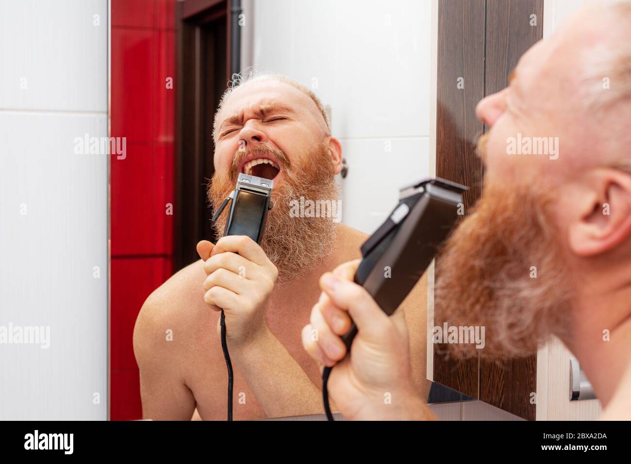 Handsome cheerful bearded man singing into hair trimmer instead of microphone in front of mirror in bathroom at home. Positive happy man joking Stock Photo