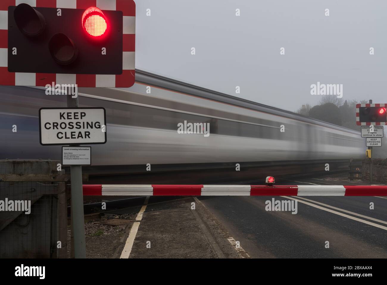 Foggy early morning commuter train traveling at speed across a road level crossing with barriers and flashing lights. Stock Photo