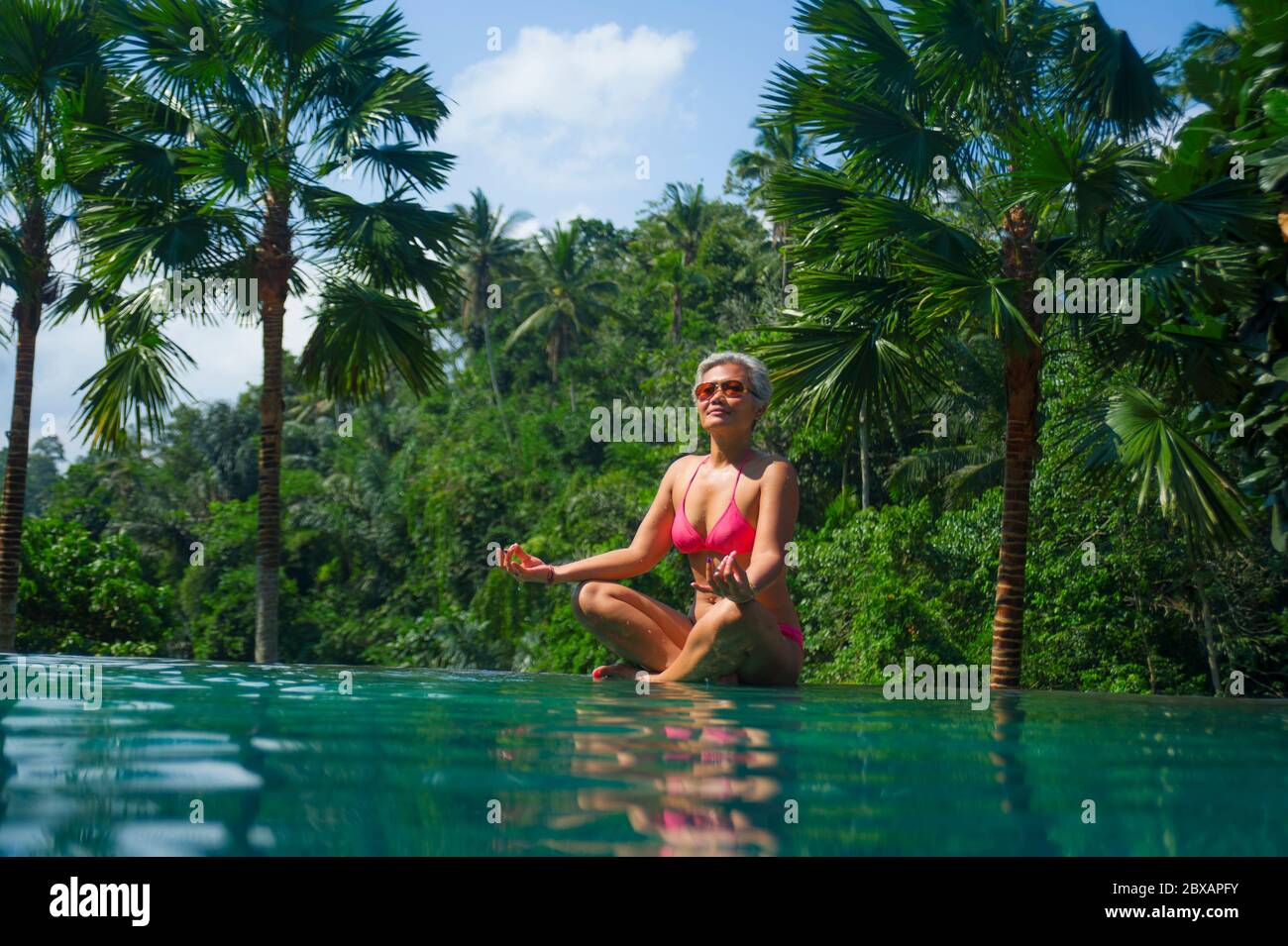 attractive and happy middle aged Asian Indonesian woman in bikini relaxed at infinity pool of exotic resort in tropical jungle sitting in lotus yoga p Stock Photo