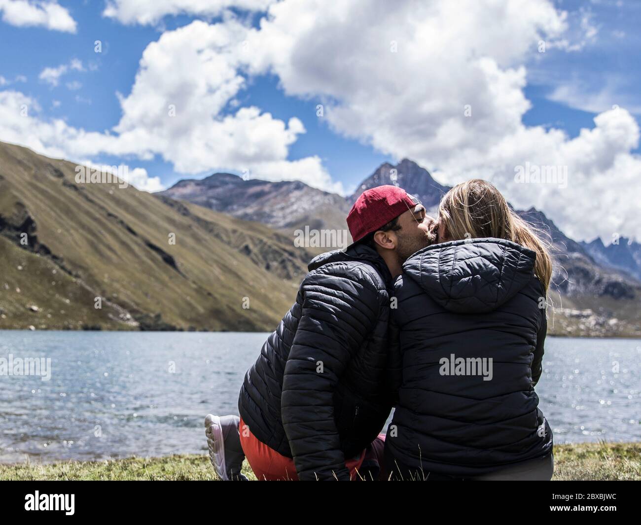 couple sitting near a lake, the boy is kissing the girl, cold, mountains, clouds and blue sky Stock Photo