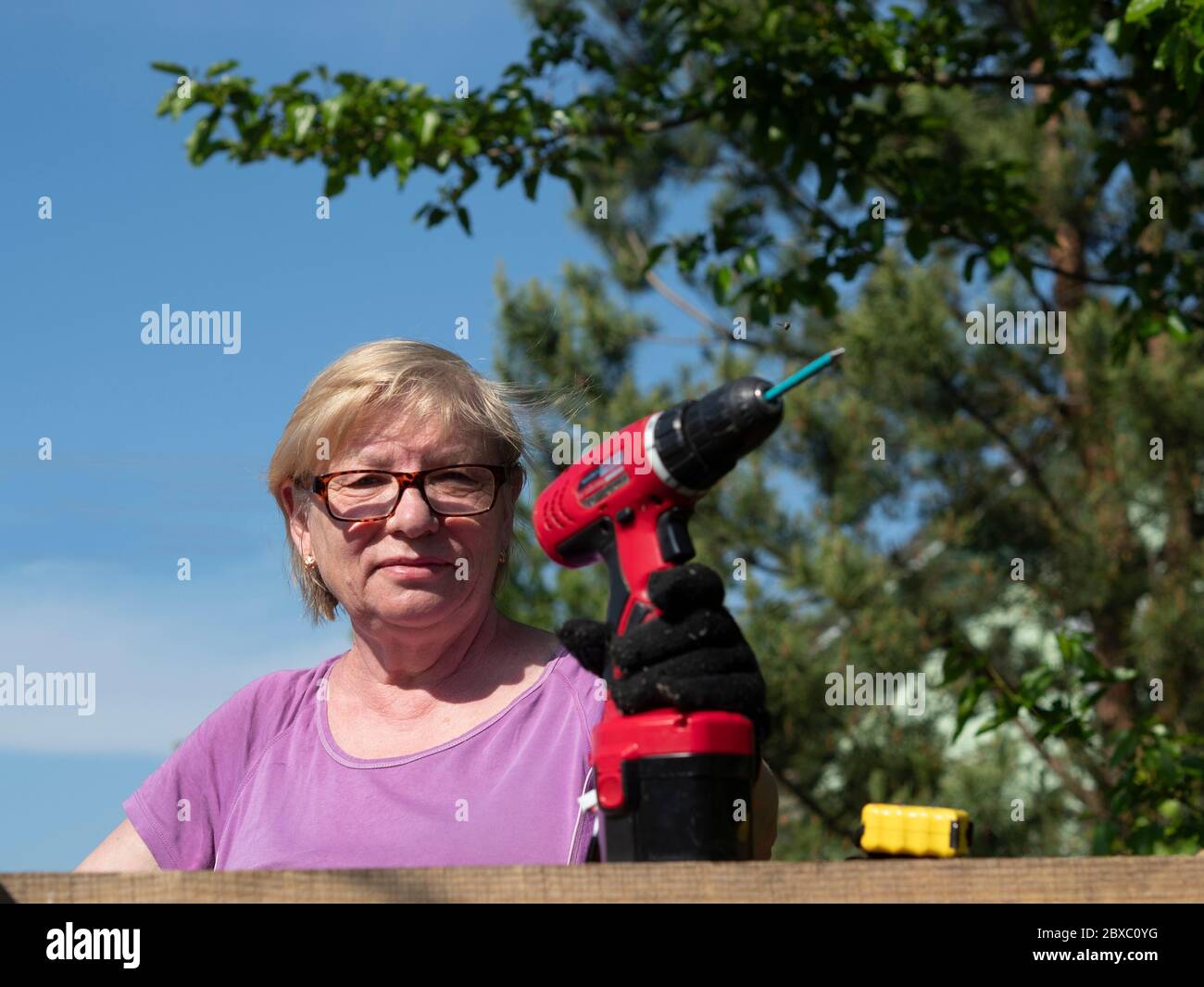 Senior Caucasian woman fastens a board on the roof of a farm building in the garden with a screwdriver Stock Photo