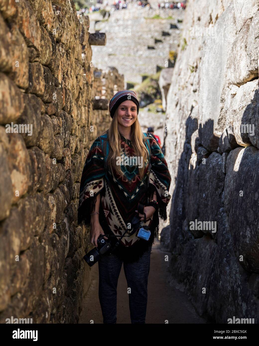 Blonde young woman standing between two stone walls with a camera in her hand and a peruvian outfit. Stock Photo