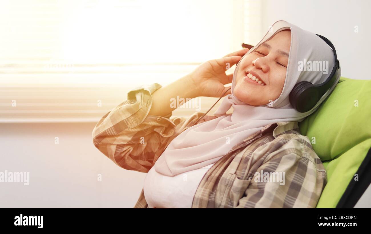 Happy young Asian muslim woman wearing hijab listening music from headphone while sitting on cozy chair, closed eyes enjoying her time Stock Photo