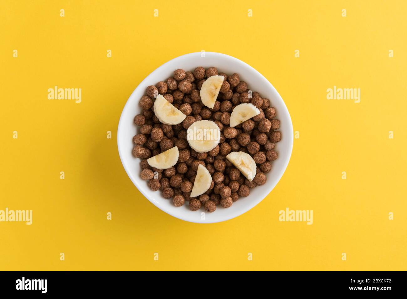 White bowl full of chocolate corn cereal balls and banana slices on yellow background, top-down view. Modern vivid breakfast image. Stock Photo