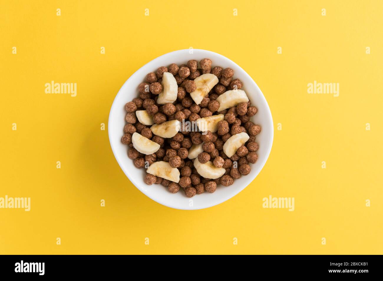 White bowl full of chocolate corn cereal balls and banana slices on yellow background, top-down view. Modern vivid breakfast image. Stock Photo