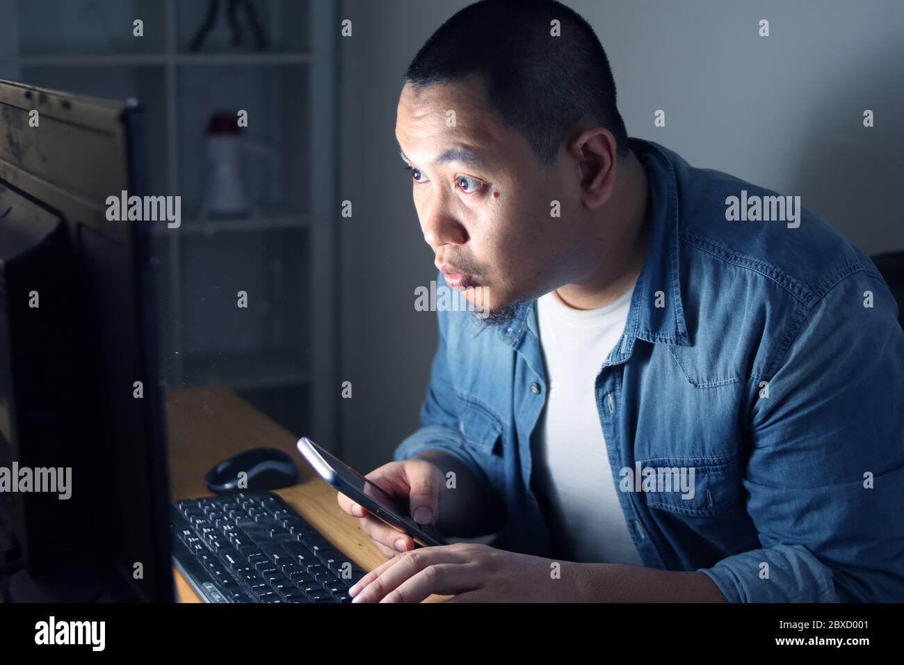 Young Asian man using smart phone and computer until late night, he is working or studying hard to reach his ambition. Student or businessman works ha Stock Photo