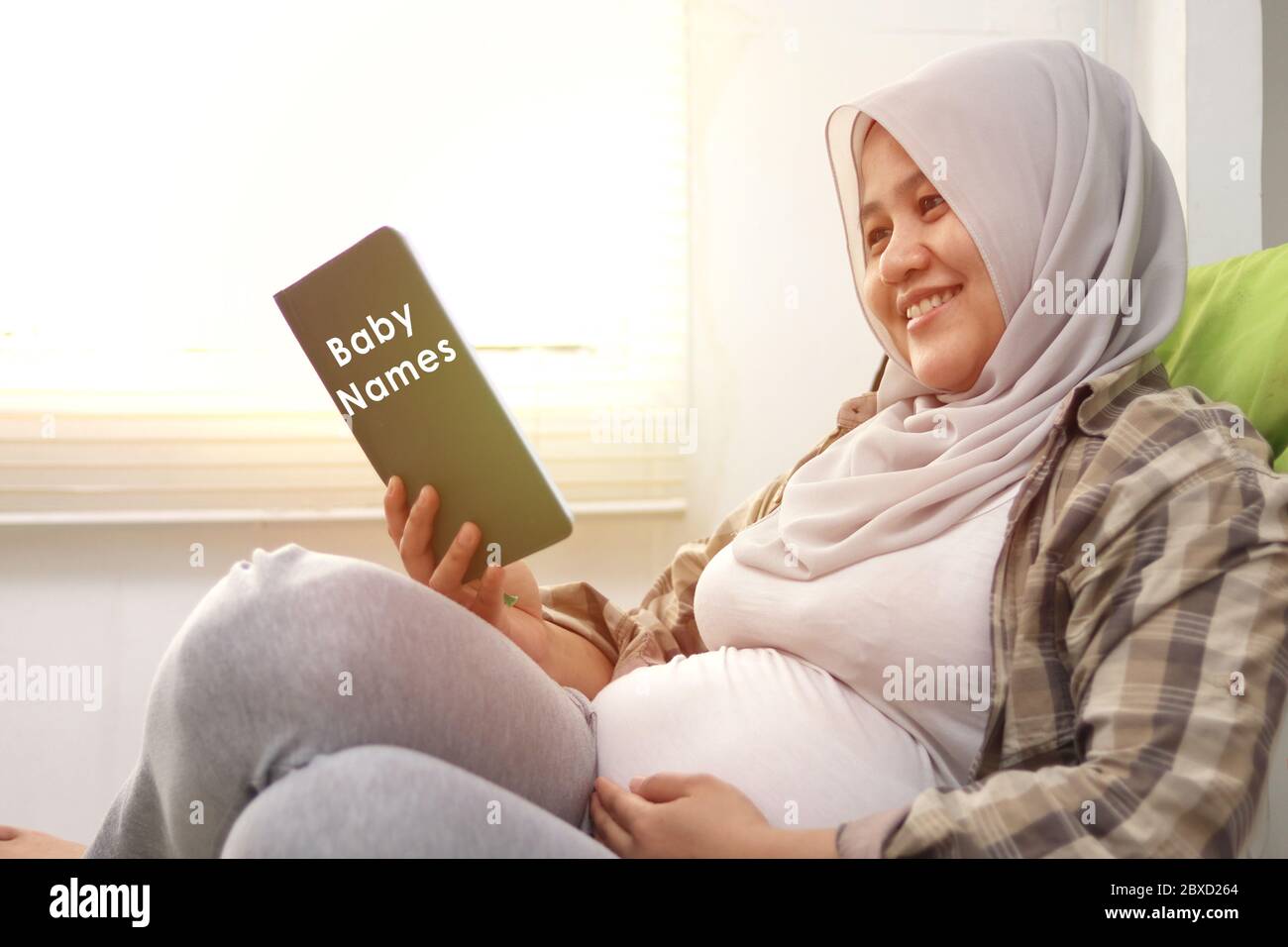 Pregnant woman sitting on couch her belly and reading baby name book. Expecting mother relaxing and smiling, preparing name for her baby Stock Photo