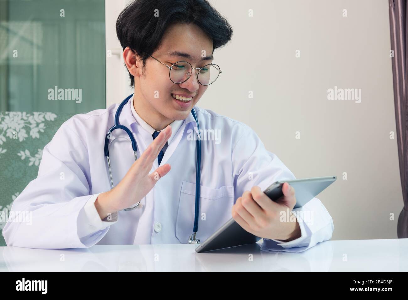 Happy Asian young doctor handsome man smile in uniform wear glasses raise hands to greet patients modern smart digital tablet computer on desk at hosp Stock Photo