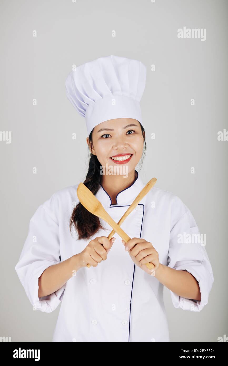 Portrait of pretty smiling young Asian cook crossing wooden spoon and fork Stock Photo