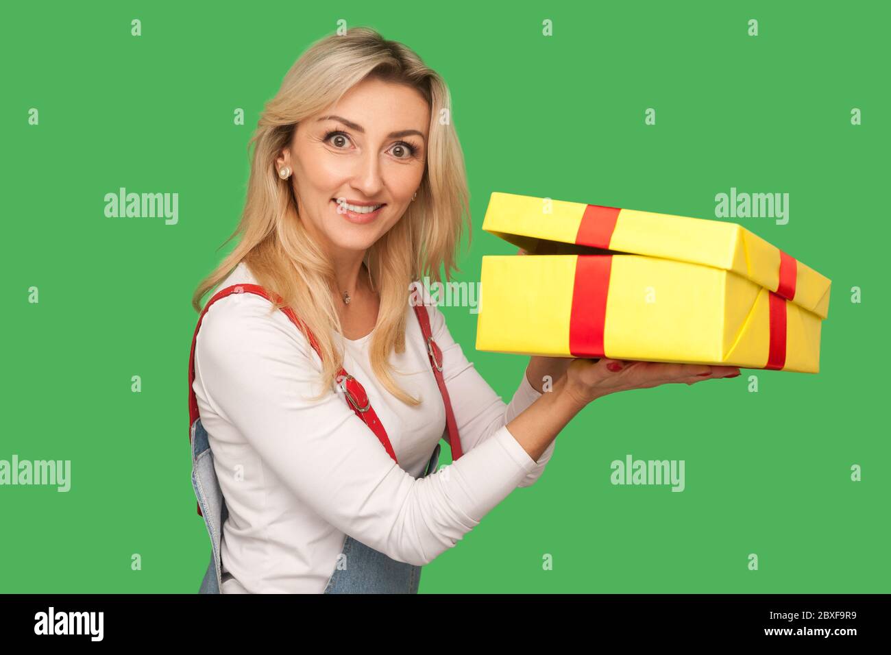 Portrait of positive pleased adult woman in denim overalls holding unwrapped gift, unpacking present and looking with toothy smile, joyful satisfied e Stock Photo