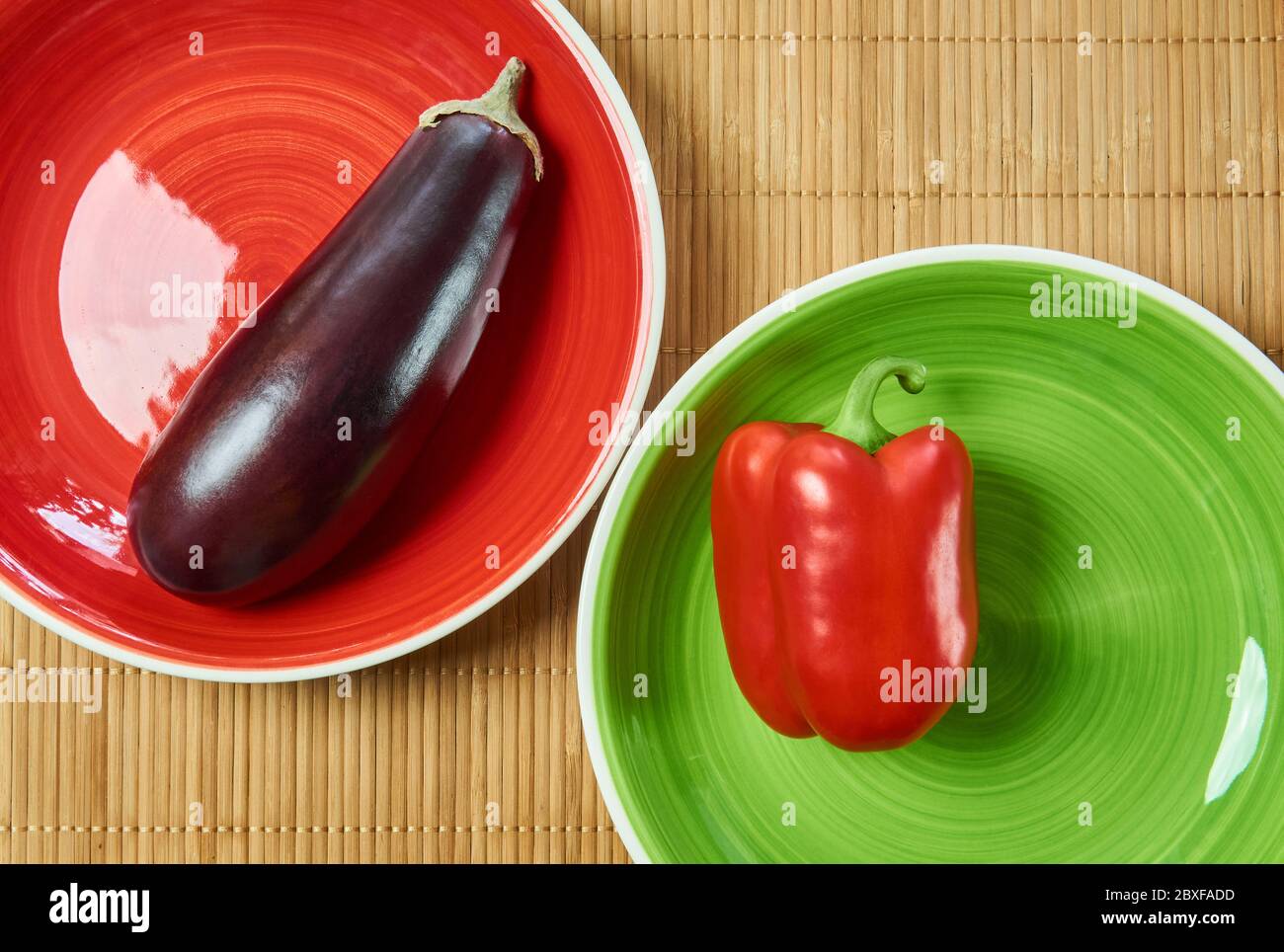 color contrasting still life - purple eggplant on a red plate and red bell pepper on a green plate next on a cane serving mat Stock Photo