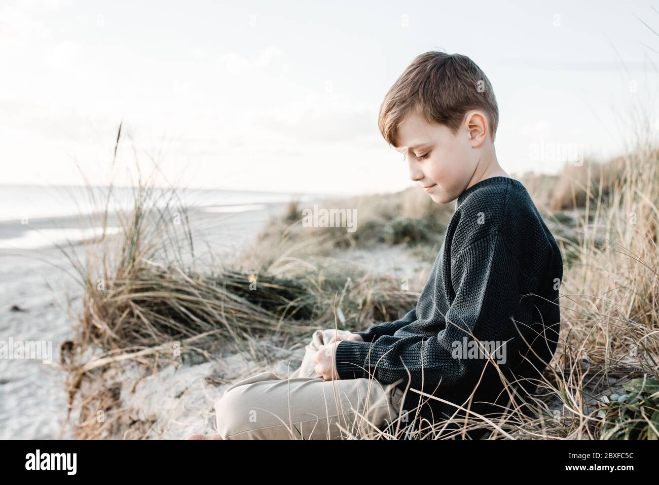 An autistic boy sitting on the grass on sand dunes at the beach in Victoria Australia Stock Photo