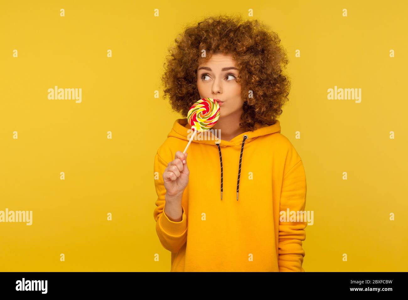 Portrait of pensive curly-haired woman in urban style hoodie licking lollipop, enjoying sweet rainbow candy and looking away with thoughtful expressio Stock Photo