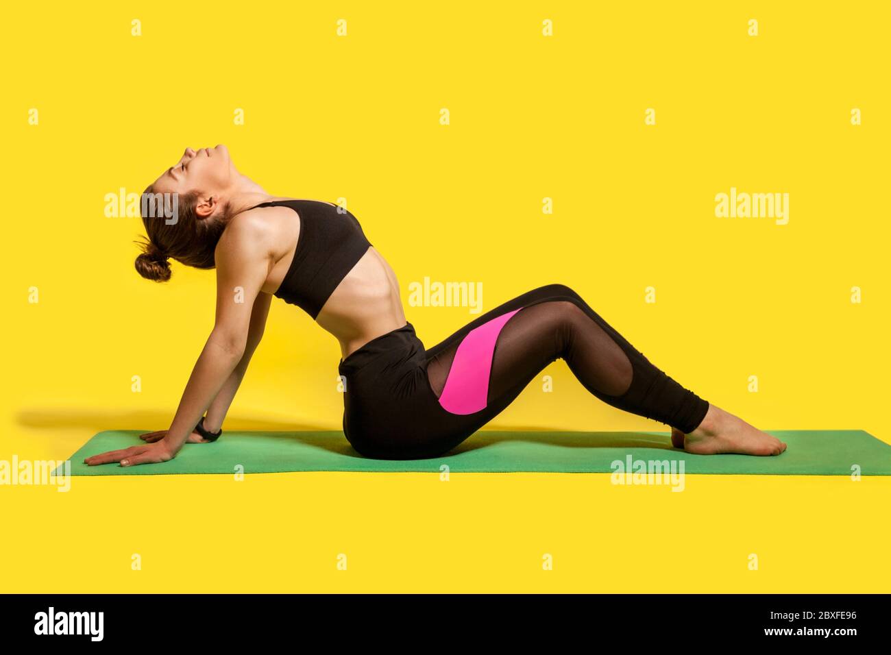 Fitness woman with hair bun in tight sportswear sitting on gym mat doing sport, bending backwards stretching spinal muscles, warming up with flexibili Stock Photo