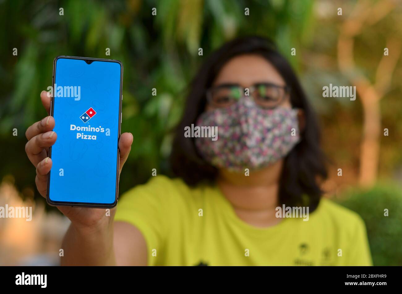 New Delhi, India, 2020. Portrait of a woman wearing a surgical mask holding a smartphone showing Domino's pizza delivery mobile app during coronavirus Stock Photo
