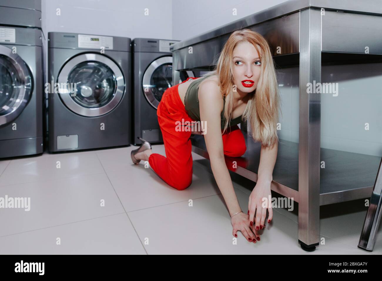 Pretty female crawls from under table in a laundry; laundry machines in background; she wears bright red trousers, high heels & a top; bright red lips Stock Photo