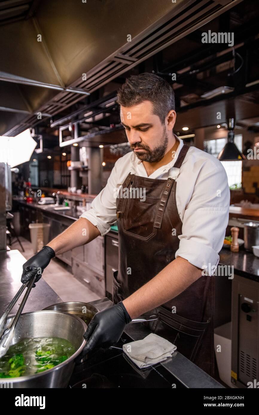 Busy bearded chef in apron standing at stove and boiling broccoli in pot at kitchen Stock Photo