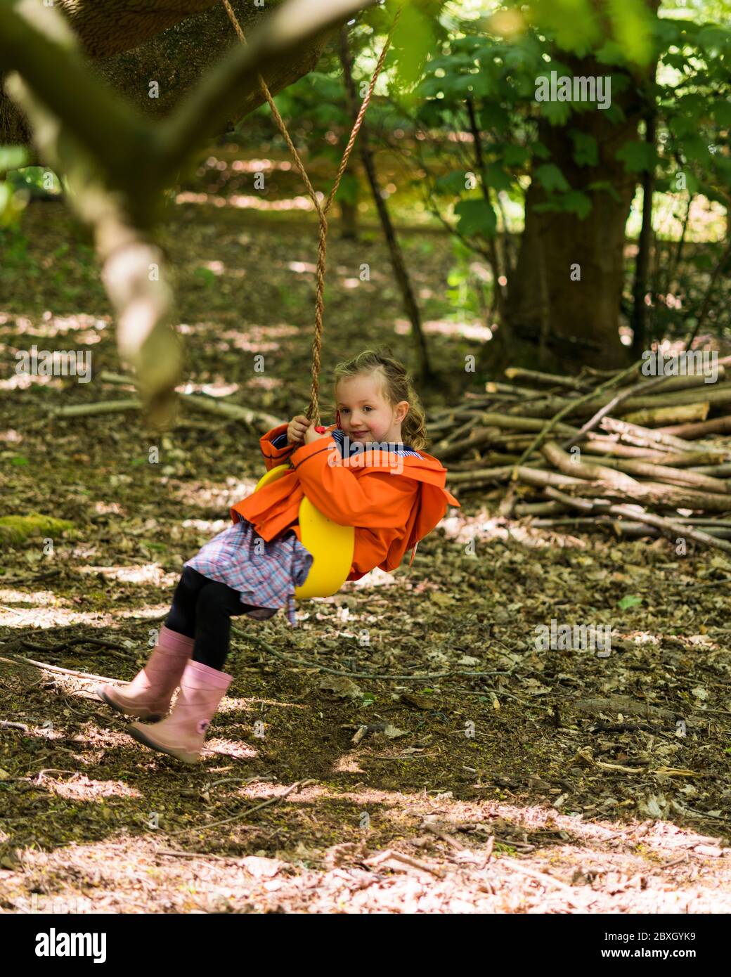 Little girl in orange raincoat playing in the woods on swing, in den, near the tree's Stock Photo