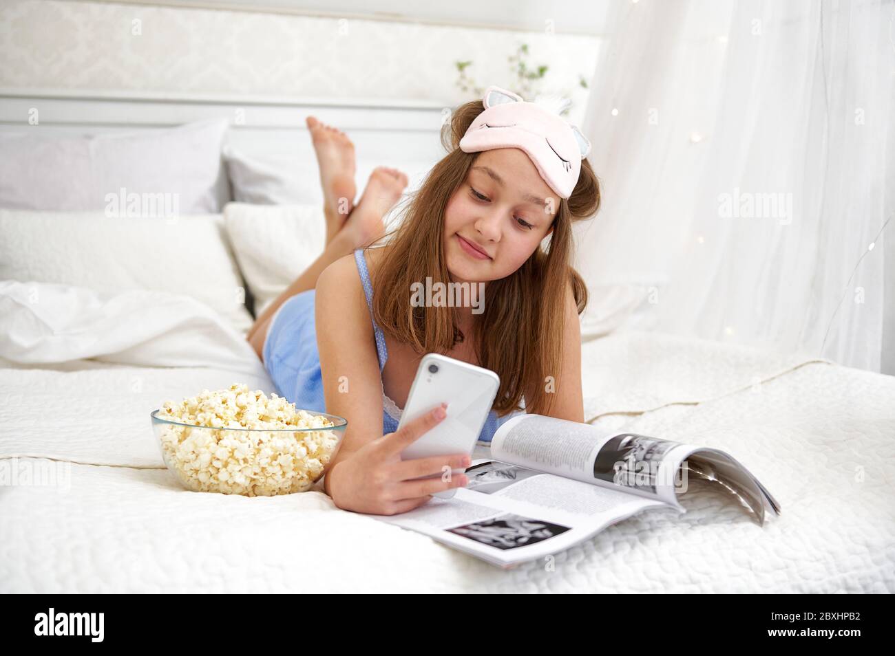 A young girl lies in bed after sleeping and browses social networks on the phone. Free time outside of school. Quarantine life. Stock Photo