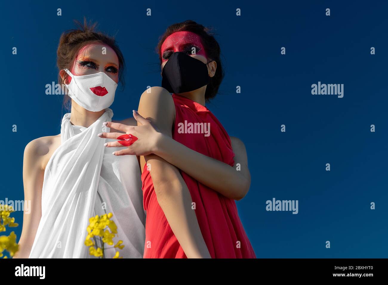 Two beautiful young brunette girls with creative bright makeup in tunics against a blue sky. One girl in a mask, the other girl closes her mouth with Stock Photo