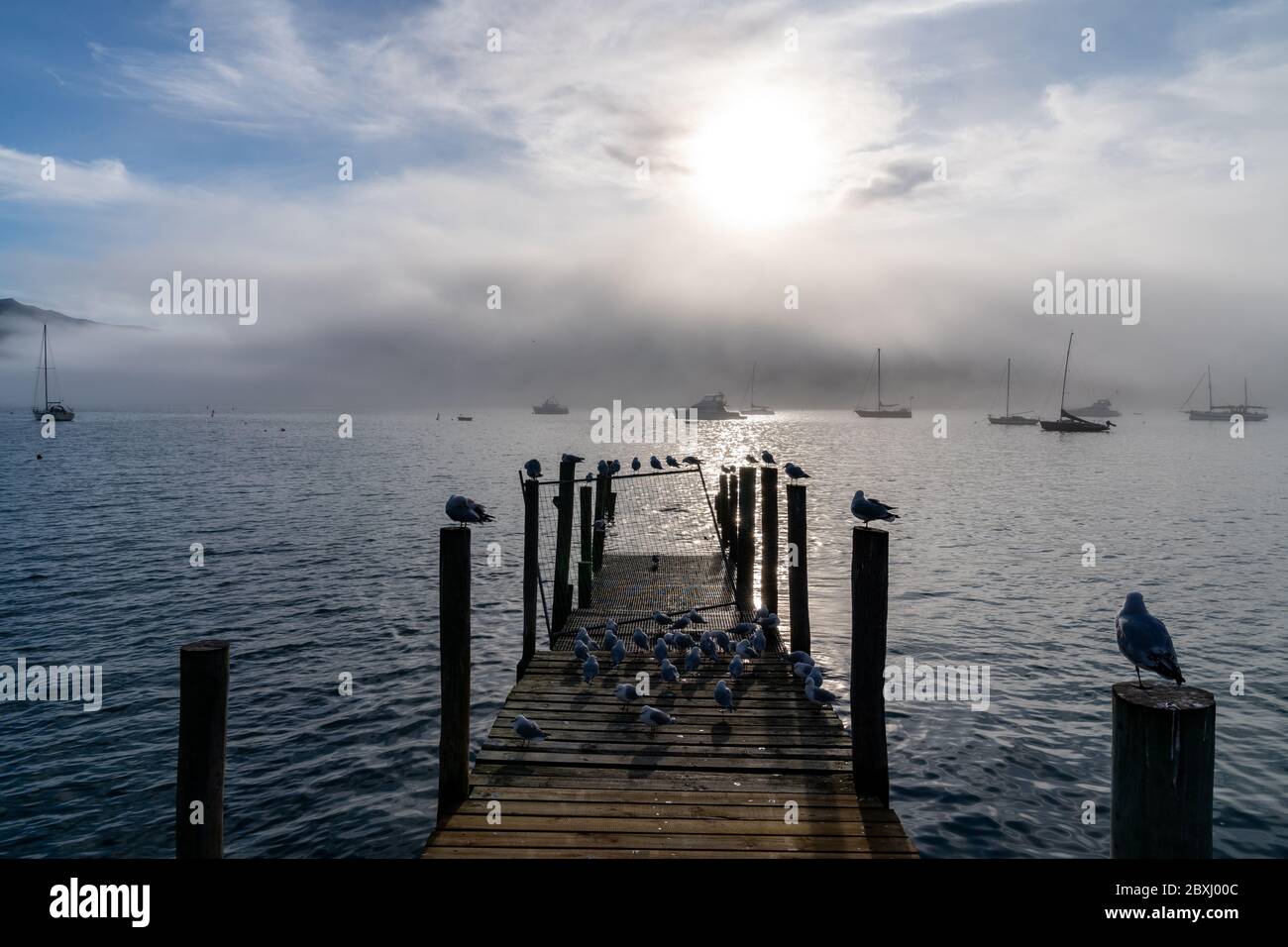 Wooden pier and sailboat at Akaroa harbor, New Zealand. Clear sky with a few white clouds and mist. Symbol for relaxation, wealth, leisure activity. P Stock Photo